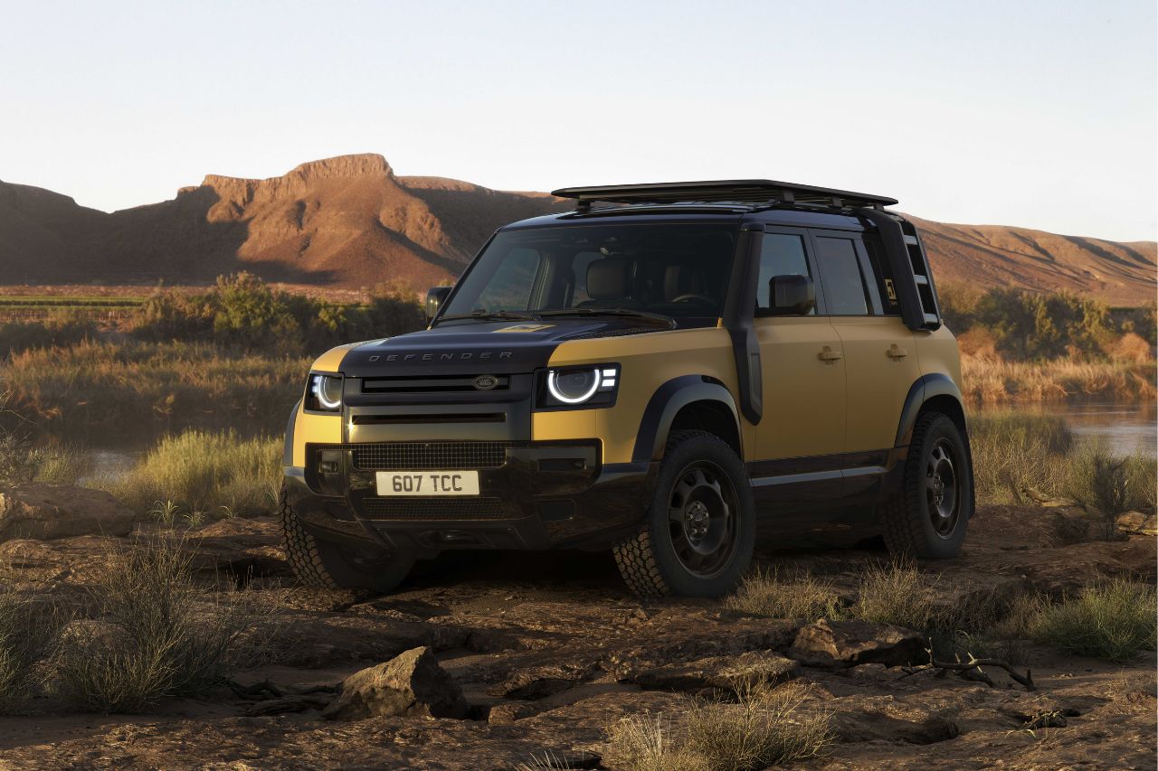 Land Rover Defender beige y negro estacionado en un terreno desértico rocoso con montañas lejanas al fondo.