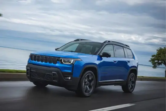 Un SUV azul de tamaño mediano circula por una carretera costera junto a una tranquila masa de agua bajo un cielo nublado.