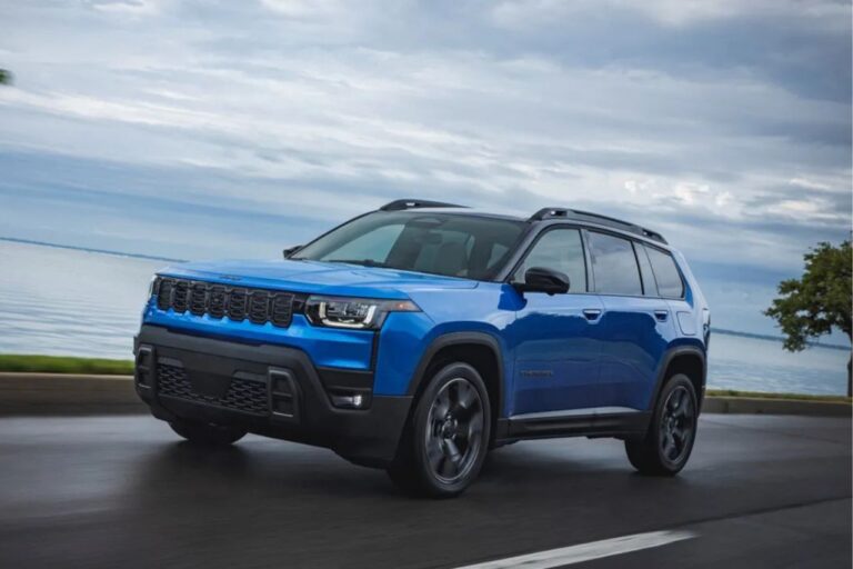 Un SUV azul de tamaño mediano circula por una carretera costera junto a una tranquila masa de agua bajo un cielo nublado.