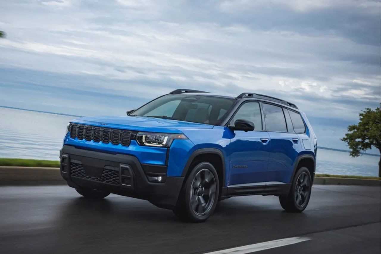 Un SUV azul de tamaño mediano circula por una carretera costera junto a una tranquila masa de agua bajo un cielo nublado.