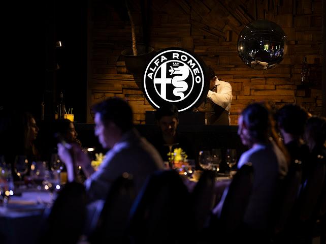 Presentación Alfa Romeo Junior San Miguel de Allende Memo Lira Dimly lit banquet with guests at tables illuminated Alfa Romeo logo on stage behind a DJ booth with a disco ball overhead