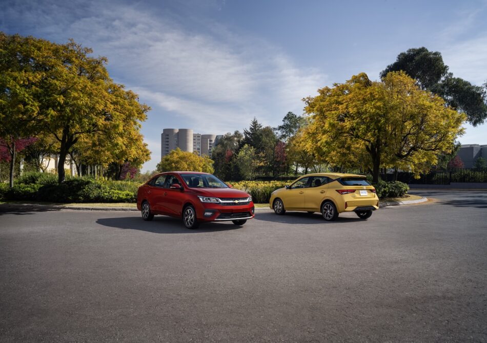 Dos coches —un sedán rojo y un utilitario amarillo— están aparcados uno frente al otro en un amplio aparcamiento asfaltado, con árboles otoñales y un cielo azul claro a sus espaldas.