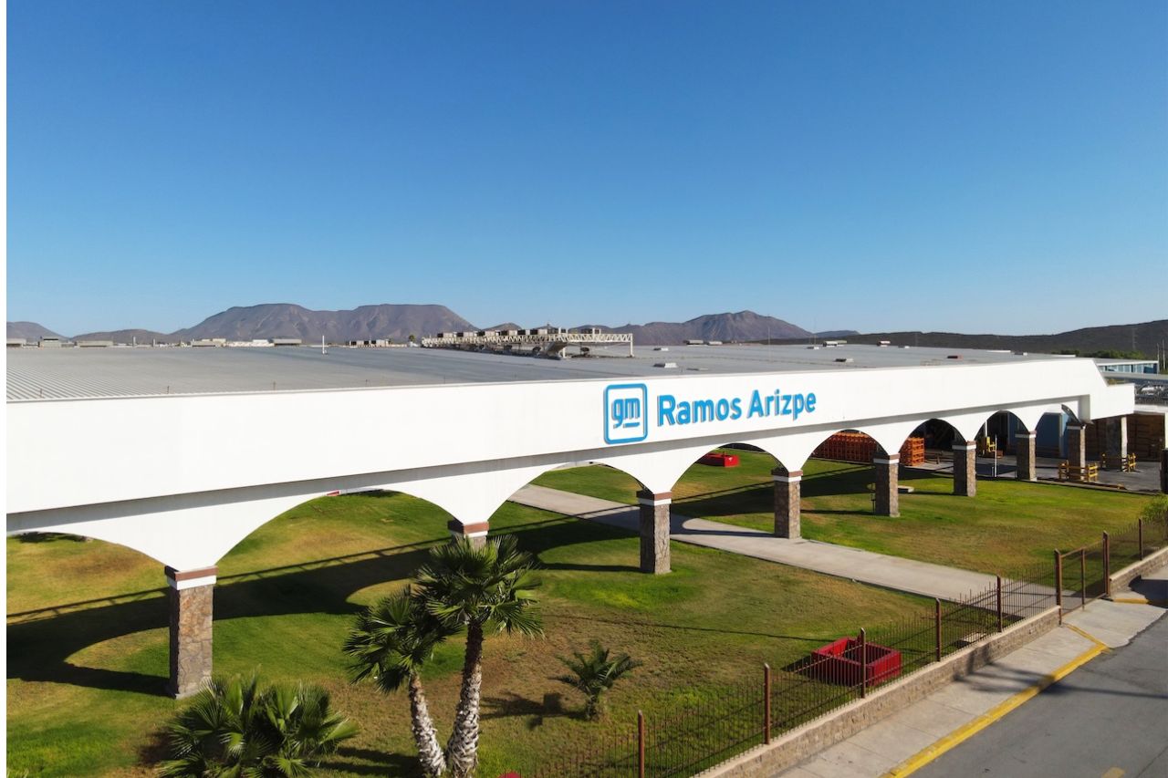 Vista exterior de unas instalaciones de GM Ramos Arizpe con una pasarela arqueada blanca, césped verde y montañas en la lejanía bajo un cielo azul y despejado.