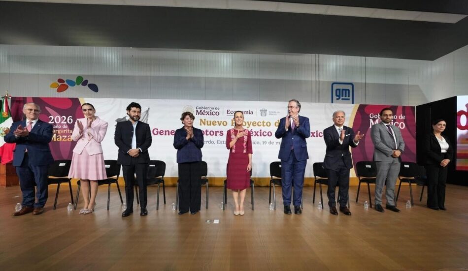 Un grupo de diez personas vestidas de manera formal, de pie en fila en un escenario, aplaudiendo, con pancartas detrás de ellas en un acto oficial.