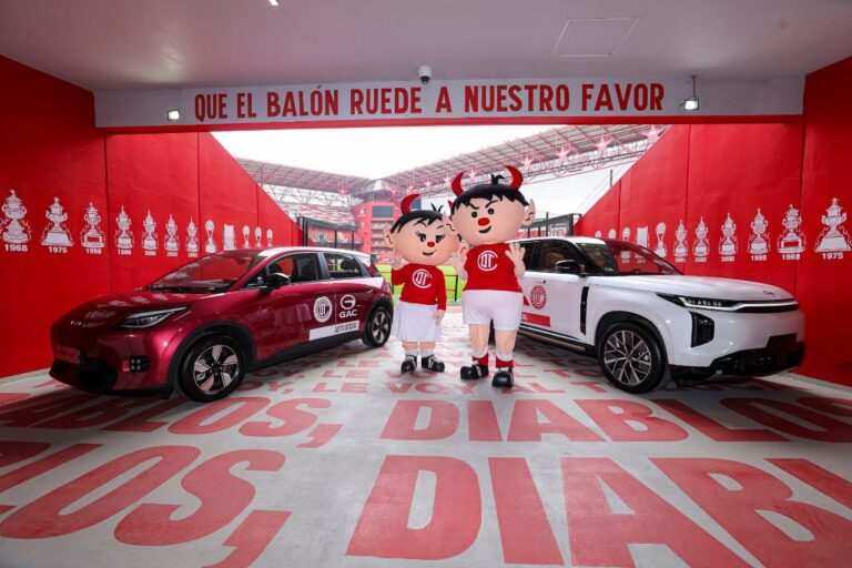 Dos mascotas disfrazadas con camisetas rojas se encuentran entre un coche rojo y un todoterreno blanco dentro de una sala de exposición de paredes rojas decorada con motivos deportivos.