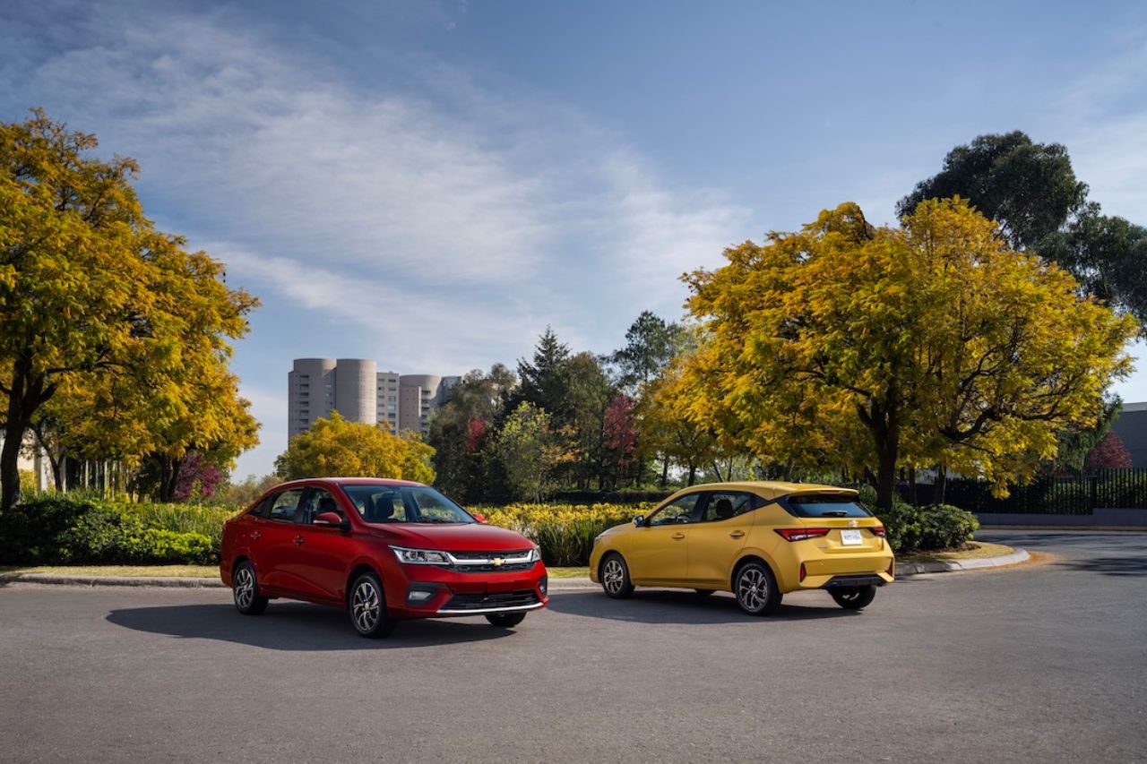 Un sedán rojo y un crossover amarillo aparcados en un aparcamiento, con árboles otoñales dorados y un cielo azul.