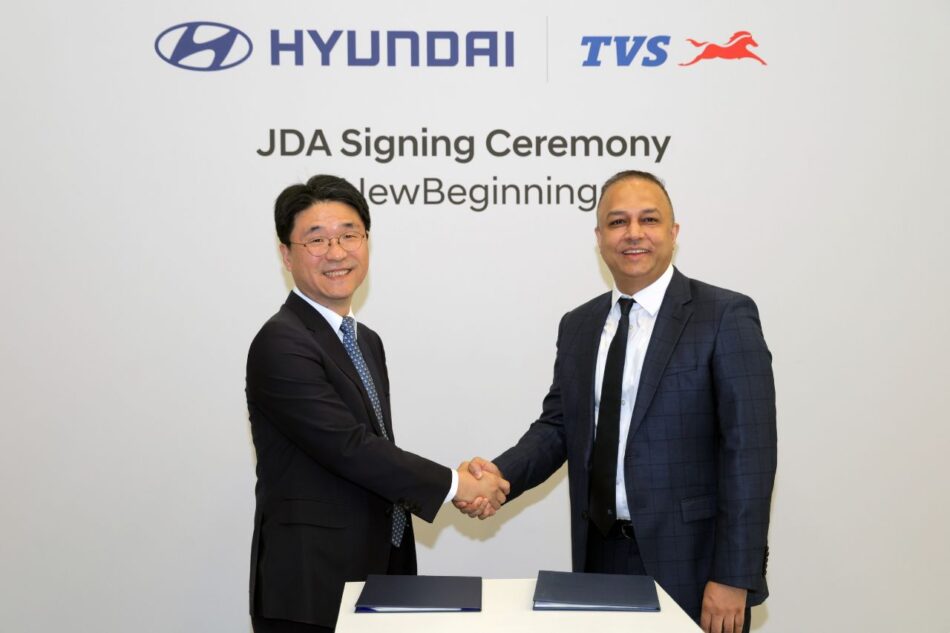 Hyundai y TVS apuestan por el futuro desarrollarán vehículos eléctricos de tres ruedas en India Memo Lira Dos hombres de traje se dan la mano en una ceremonia de firma con los logotipos de Hyundai y TVS encima y el texto Ceremonia de firma JDA en la pared detrás de ellos