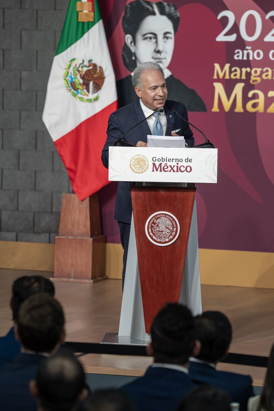 Paco Garza pronunciando un discurso en un estrado con el escudo de México y la bandera mexicana al fondo