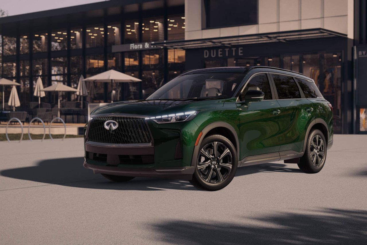 Un SUV Infiniti verde aparcado frente a un edificio moderno, con una terraza y sombrillas al fondo.