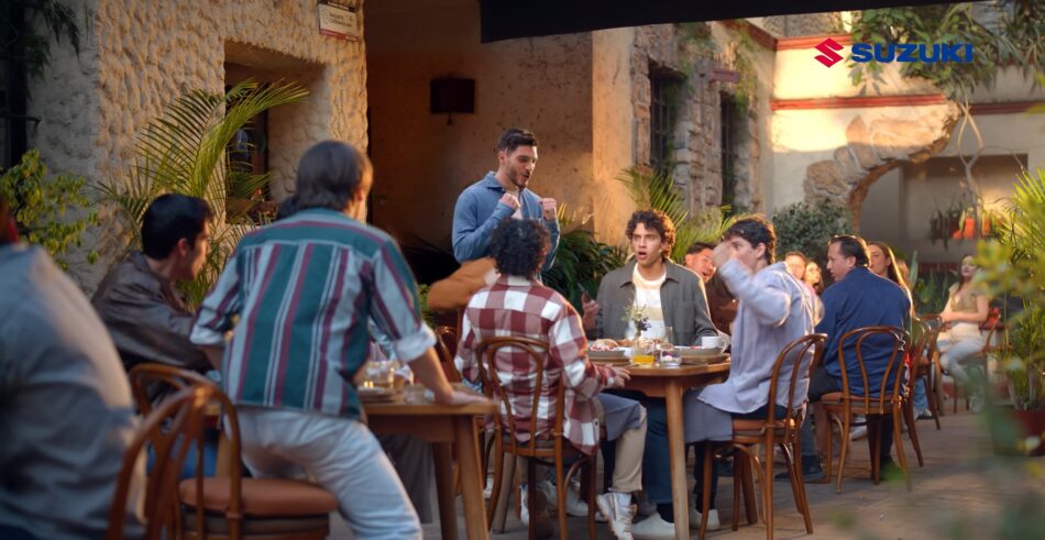 Suzuki presenta nueva campaña con Raúl Jiménez y una narrativa más humana Memo Lira Escena de un restaurante al aire libre con gente sentada en las mesas un hombre está de pie y gesticula mientras los demás le escuchan se ve el logotipo de Suzuki en la esquina superior derecha