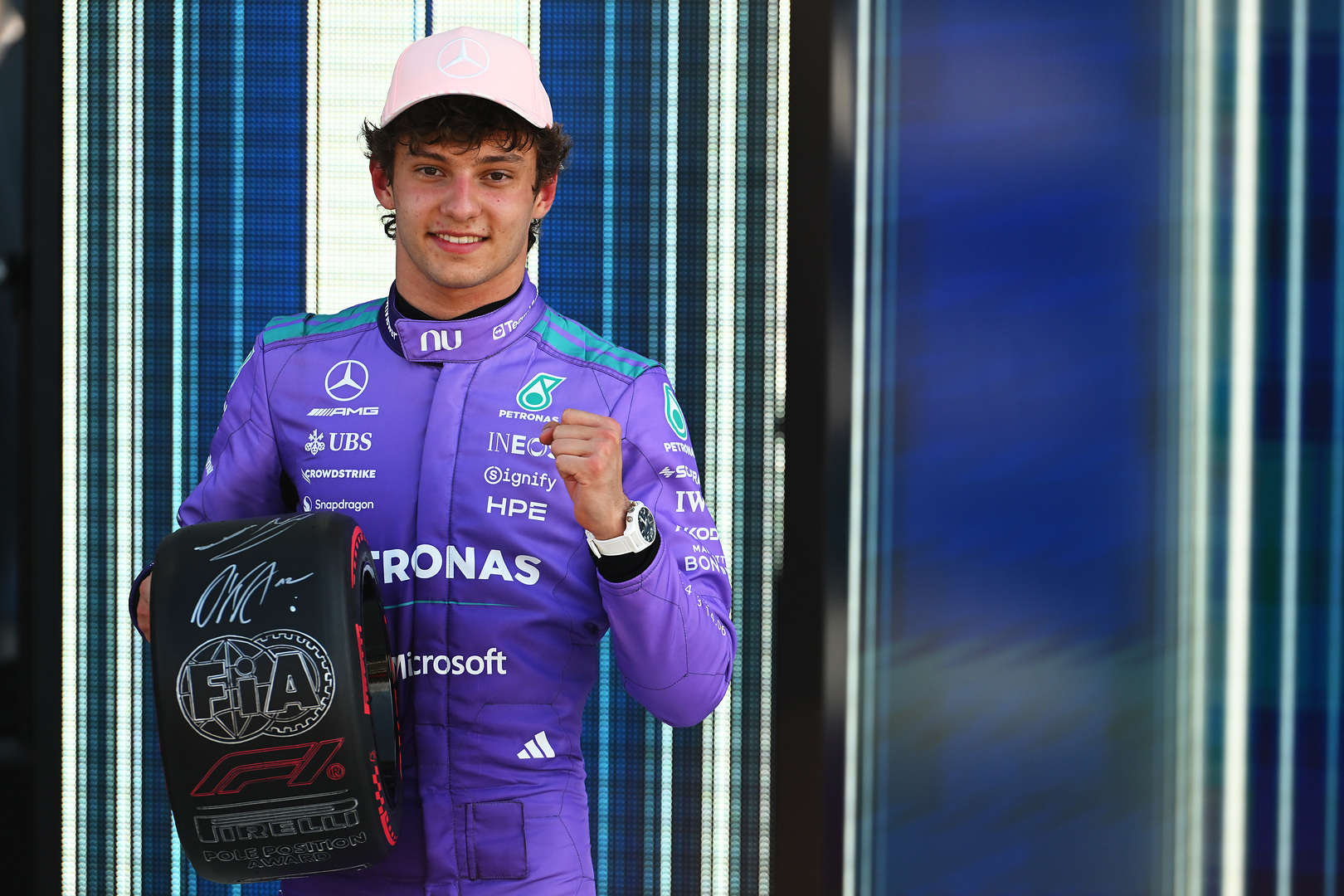 Formula 1 driver in a purple Mercedes racing suit and pink cap poses with a signed FIA F1 helmet.
