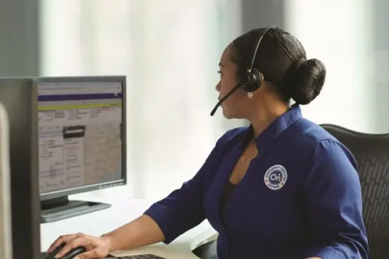 Agente de atención al cliente vestido con un uniforme azul y auriculares, trabajando en un escritorio con un ordenador y un monitor cerca.
