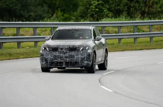Camouflaged prototype SUV driving on a road with a metal guardrail, driver visible through the windshield.