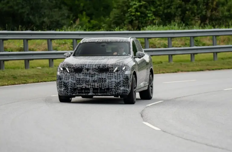 Camouflaged prototype SUV driving on a road with a metal guardrail, driver visible through the windshield.