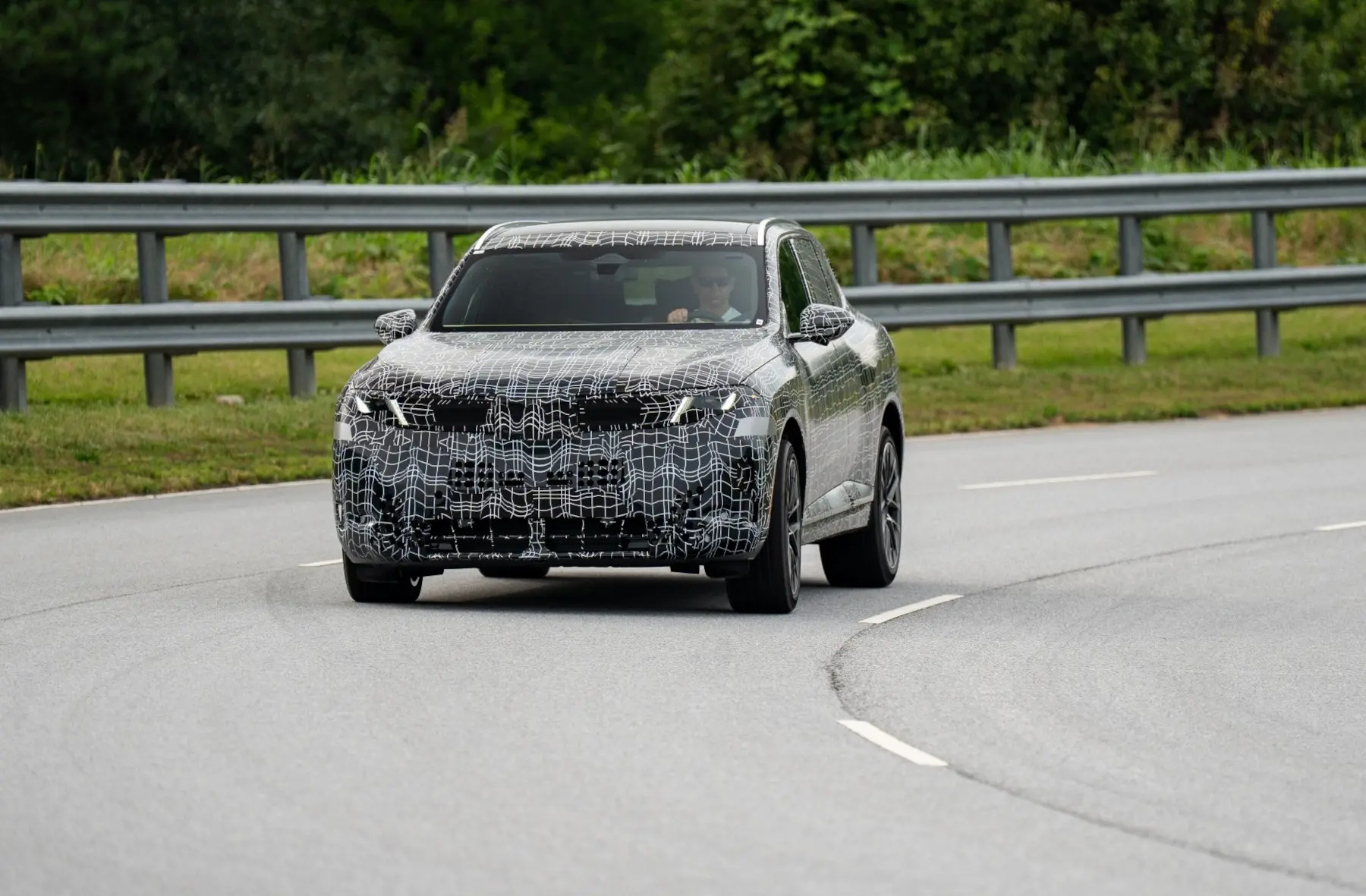 Camouflaged prototype SUV driving on a road with a metal guardrail, driver visible through the windshield.