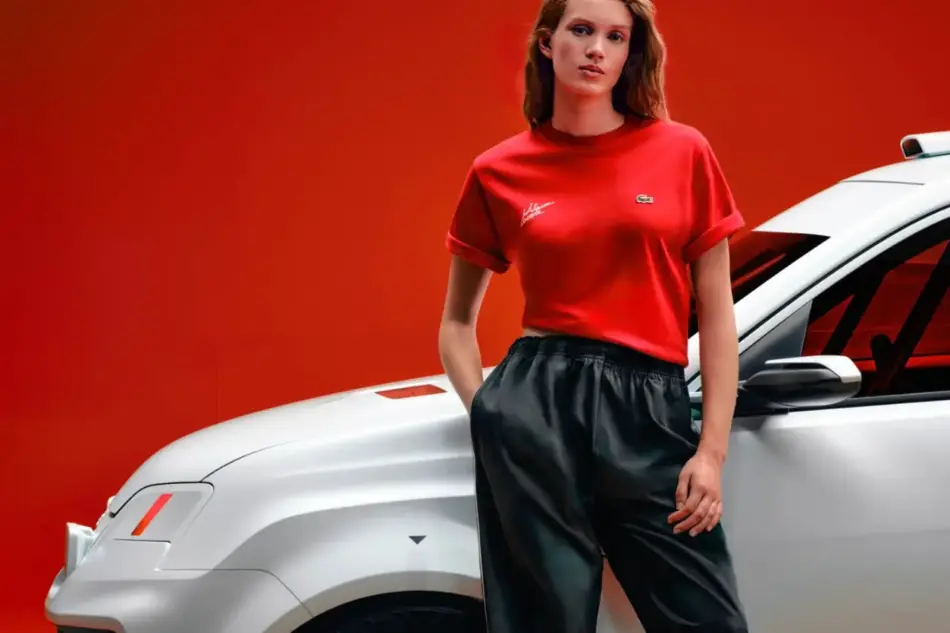 Woman in a red t shirt and black pants stands beside a white car against a red backdrop posing confidently for an ad