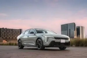 Silver Kia K4 GT-Line sedan parked on a paved plaza with modern office buildings in the background at sunset.