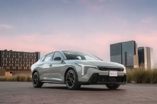Silver Kia K4 GT-Line sedan parked on a paved plaza with modern office buildings in the background at sunset.