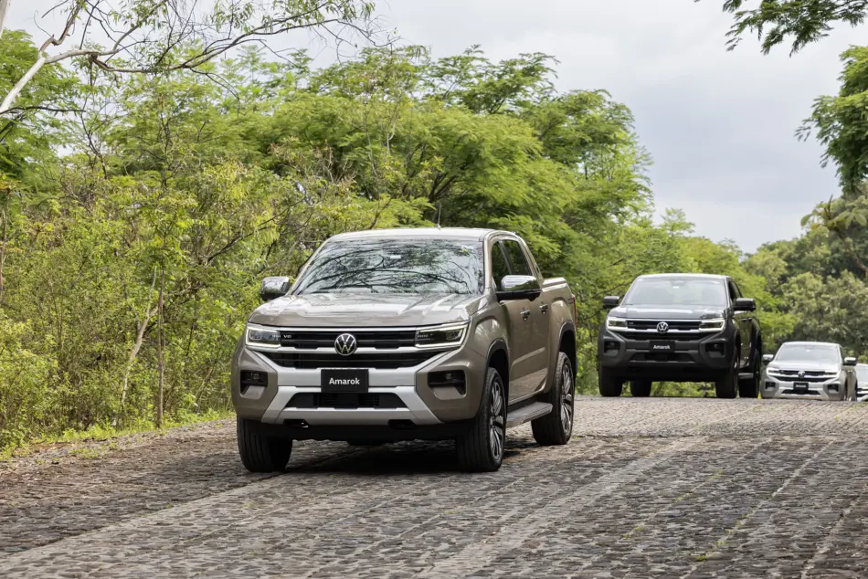 Volkswagen Amarok 2026 diferentes versiones Diseño exterior Memo Lira Several Volkswagen Amarok pickup trucks drive along a cobblestone road with lush green trees on both sides the lead car in front
