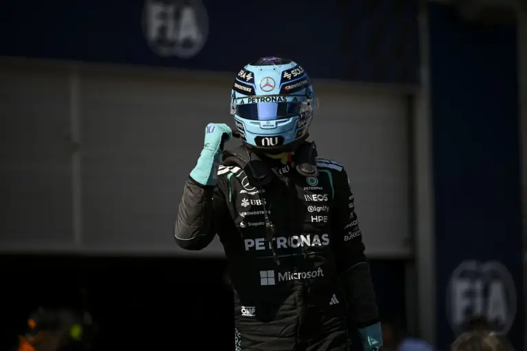 Formula 1 driver in racing suit raises a gloved fist, wearing a turquoise helmet with sponsor logos (Petronas, Mercedes) and a black team suit with Microsoft logo visible, in a dim pit area.