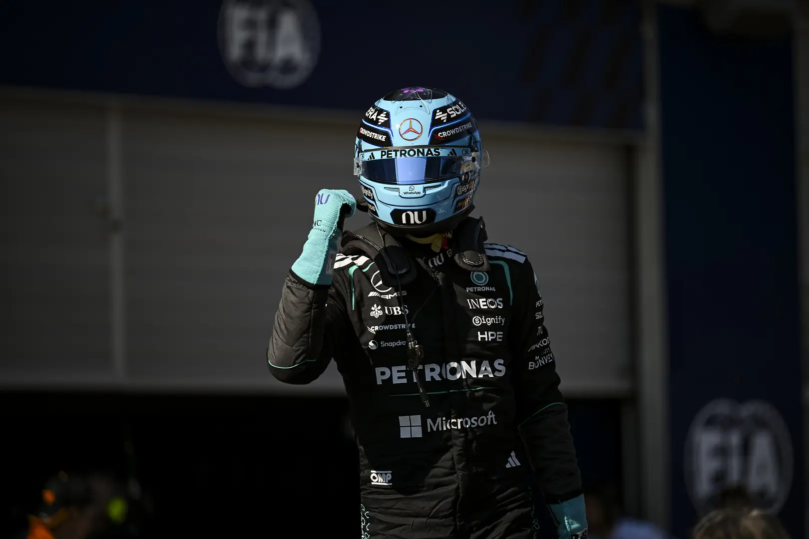 Formula 1 driver in racing suit raises a gloved fist, wearing a turquoise helmet with sponsor logos (Petronas, Mercedes) and a black team suit with Microsoft logo visible, in a dim pit area.