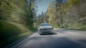 Camouflaged sedan driving on a two-lane road through a forest, motion blur creating a speed effect on both sides of the trees and guardrails