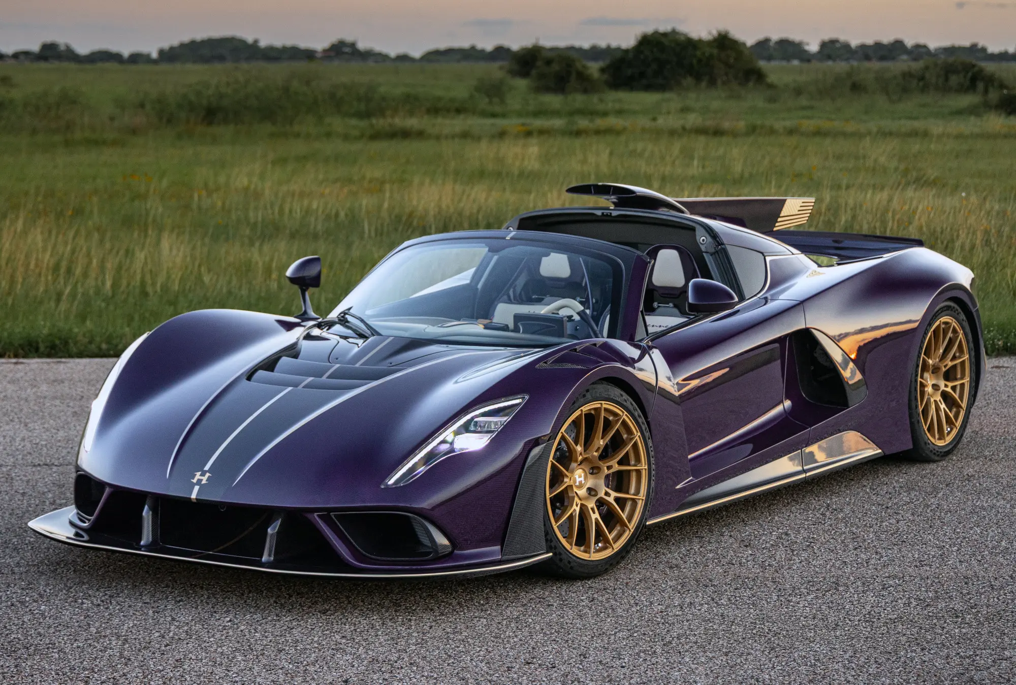 Purple convertible sports car with gold wheels on a paved road, open cockpit, field backdrop at sunset.