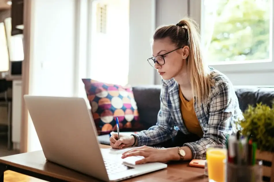 Generación Z en computadora Memo Lira Una mujer con gafas está sentada ante un escritorio de madera escribiendo en un papel junto a un ordenador portátil abierto en un salón muy luminoso